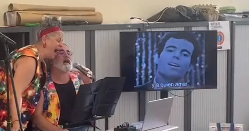 Angie y Albert, con camisas de colores, cantan abrazados junto a una pantalla con un vídeo de Julio Iglesias.
