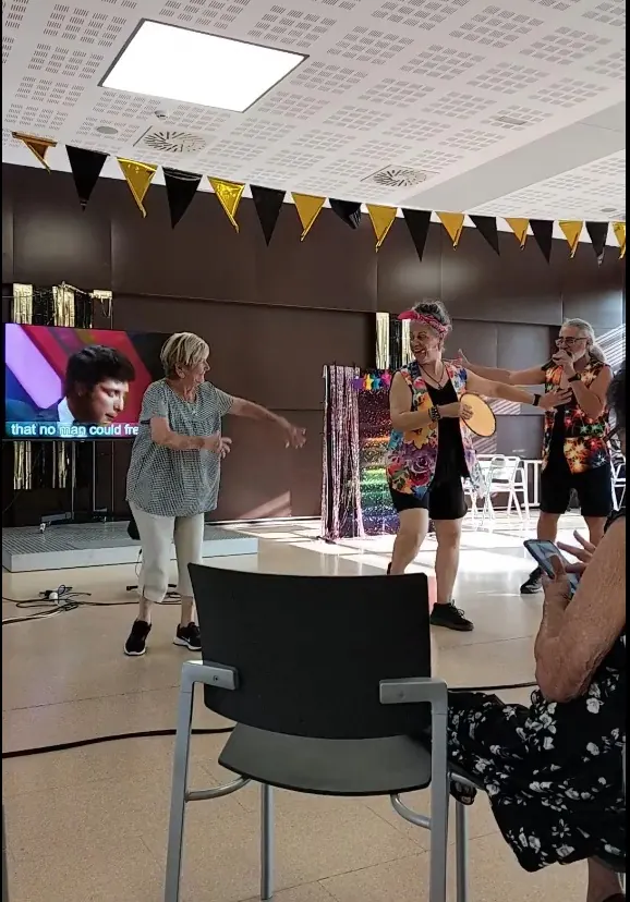 Público sentado escuchando a Angie y Albert, que cantan con los brazos en alto frente a la pantalla.